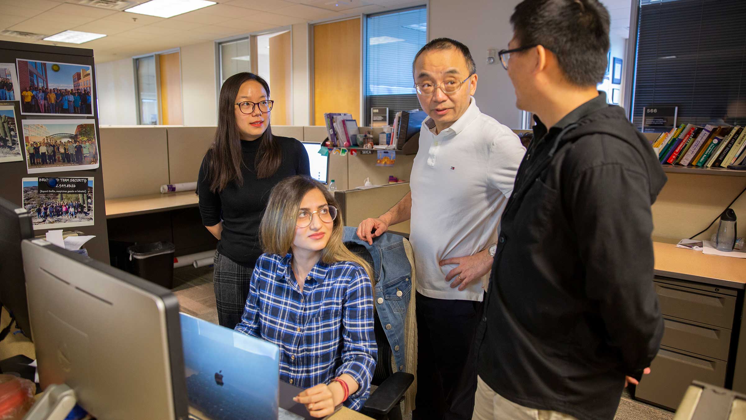 A faculty memeber stands at a desk with three students, discussing data on the computer screen