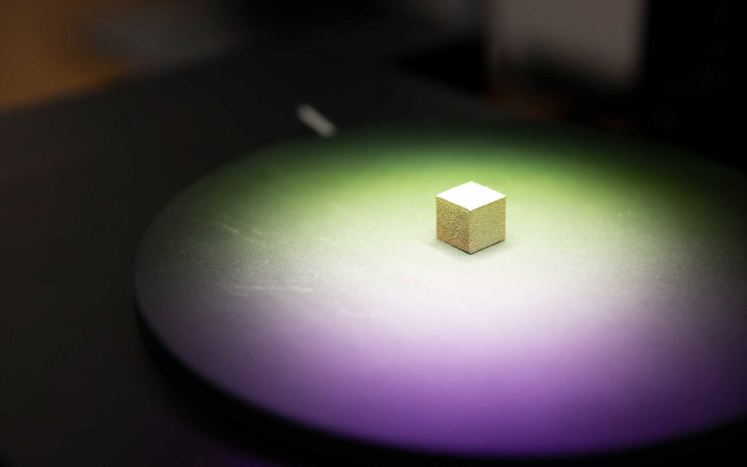 A small cube of manufactured metal is spotlighted with purple and green on a table