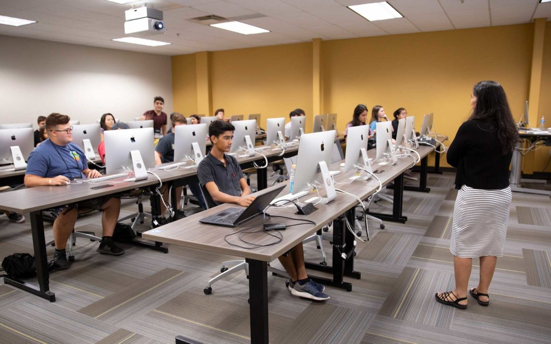 A group of students sit in a computer lab while their professor teaches at the front