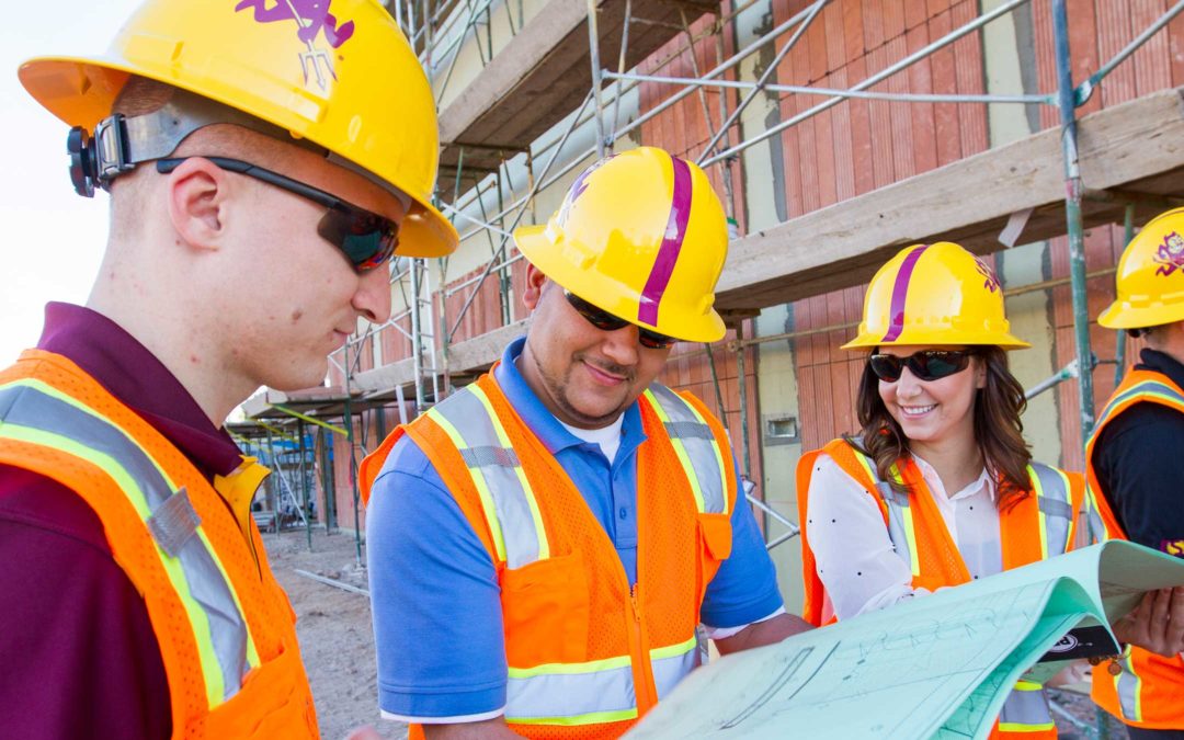 A group of students in hardhats stand at a construction site reviewing planning documents