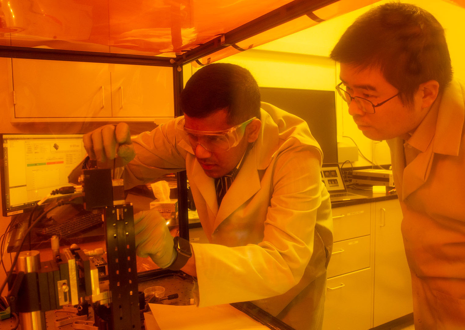 Two researchers look at a piece of lab equipment in a research lab under yellow lights.