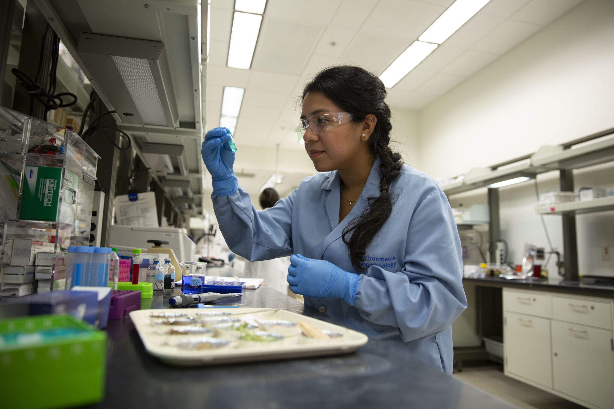 A female PhD student, dressed in lab gear, observes an object closely in her hand while in her lab.